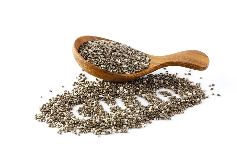 Chia seeds spilling from a wooden spoon onto a pile of chia seeds on a white background