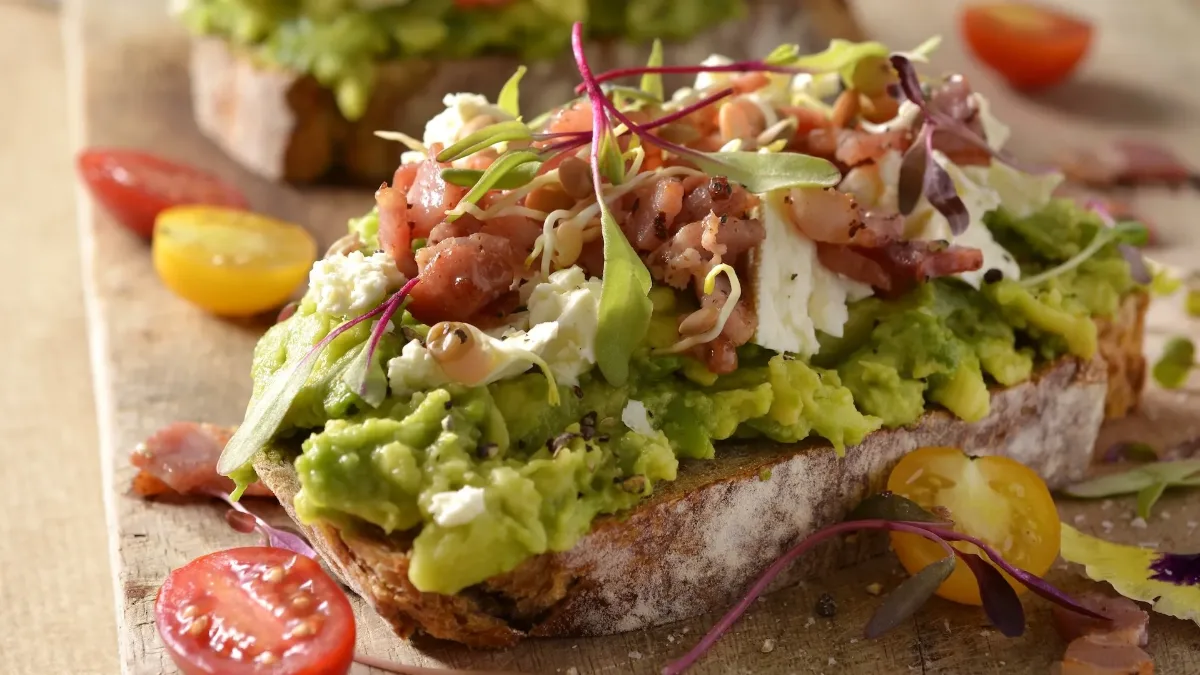 An open sourdough sandwich topped with mashed avocado, feta, bacon and micro herbs, with cherry tomatoes alongside
